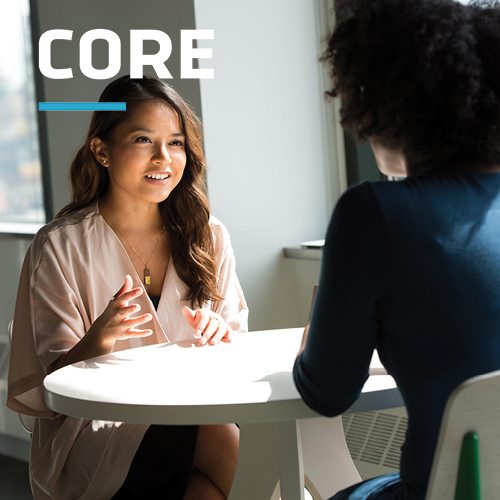 Two women sit at a round table in a bright, modern office, engaged in a professional discussion. The "CORE" branding is positioned in the top left corner, highlighting the context of structured interviews or assessments.