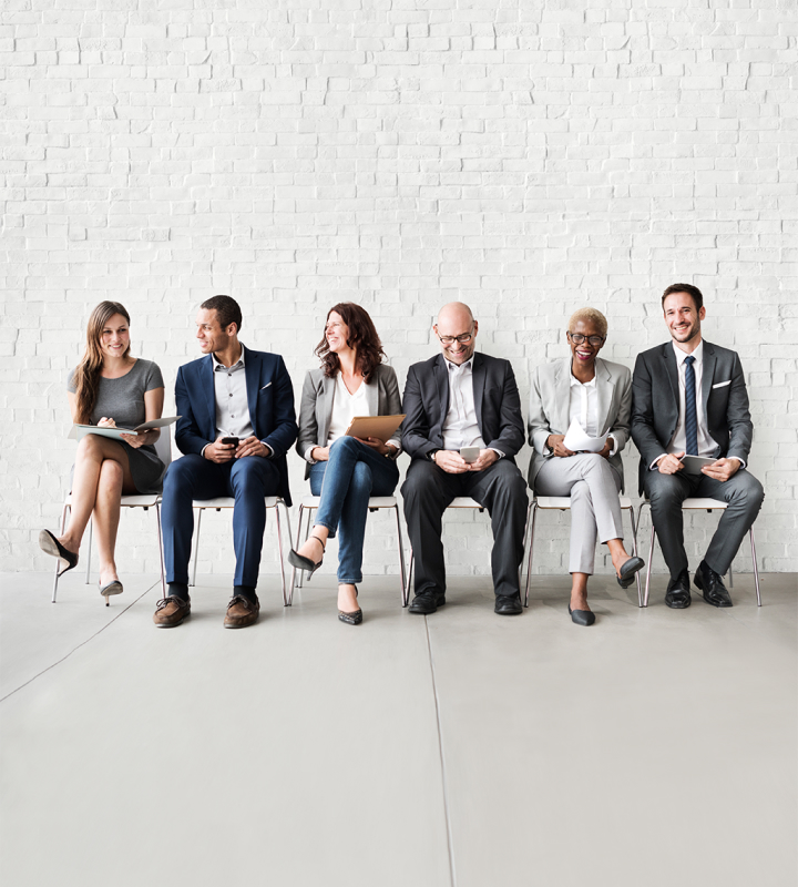 A diverse group of six professionals sitting in a row against a white brick wall, engaged in conversation and smiling, representing an inclusive and dynamic workplace environment.